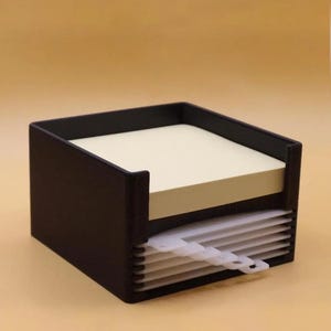 May include: A black square desk organiser featuring a top compartment filled with a stack of pale yellow square sticky notes and a lower section containing a series of white plastic dividers. The text HUMAN MADE is printed in the bottom right corner.