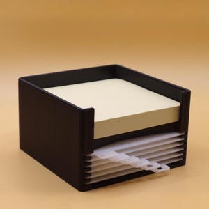 May include: A black square desk organizer featuring a top compartment filled with a stack of pale yellow square sticky notes and a lower section containing a series of thin, white, semi-transparent plastic dividers with small tabbed edges.