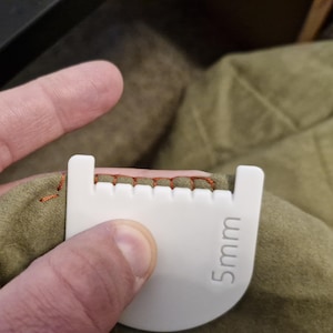 May include: A white plastic sewing spacing tool with a curved edge and notched teeth, featuring the text 5mm printed on the surface. The tool is positioned against a piece of olive green fabric with orange thread stitching along the edge.