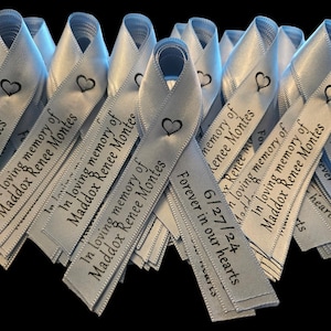 May include: A stack of light blue satin ribbons with the text "In loving memory of Maddox Renee Montes" and "Forever in our hearts 6/27/24" printed on them.