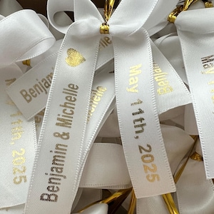 May include: White satin ribbon bows with gold lettering. The ribbon says "Benjamin & Michelle" and "May 14th, 2025".