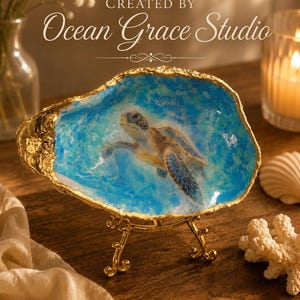May include: A decorative oyster shell dish featuring a painted sea turtle swimming in blue water, with gold-painted edges, displayed on a small gold metal stand. The text "CREATED BY Ocean Grace Studio" and the URL "https://oceangracestudio.etsy.com" are visible.
