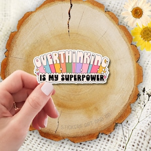 May include: A colourful sticker with the text "OVERTHINKING IS MY SUPERPOWER" in a retro font. The sticker has a white border and features a rainbow design with stars and flowers. The sticker is held by a hand on a wooden surface.