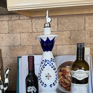 May include: A white ceramic olive oil dispenser with blue and green floral designs, a black glass bottle of olive oil labeled "Viva Oliva", and a brown glass bottle of olive oil labeled "Olio Extra Vergine di Oliva" on a black slate surface.