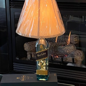 May include: A lamp made from a clear glass bottle with a gold label that reads "Johnnie Walker Blue Label" and a white lampshade. The bottle is lit from inside with white fairy lights. The lamp is sitting on a black box with the same label as the bottle.