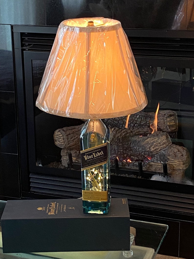 May include: A lamp made from a clear glass bottle with a gold label that reads "Johnnie Walker Blue Label" and a white lampshade. The bottle is lit from inside with white fairy lights. The lamp is sitting on a black box with the same label as the bottle.