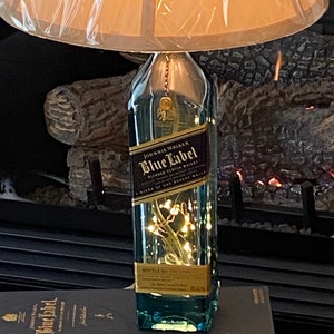 May include: A clear glass bottle lamp with a brown lampshade. The bottle is decorated with a gold label that reads "Johnnie Walker Blue Label Blended Scotch Whisky" and has a string of white lights inside. The bottle is sitting on a black box with a gold label that reads "Johnnie Walker Blue Label".