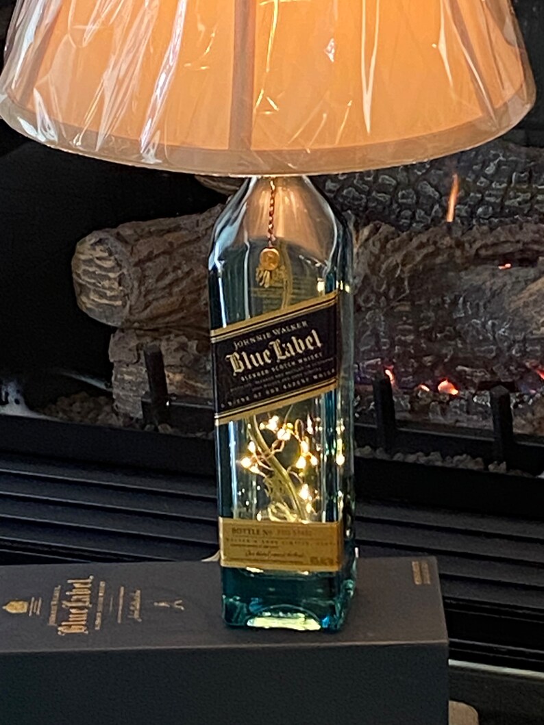 May include: A clear glass bottle lamp with a brown lampshade. The bottle is decorated with a gold label that reads "Johnnie Walker Blue Label Blended Scotch Whisky" and has a string of white lights inside. The bottle is sitting on a black box with a gold label that reads "Johnnie Walker Blue Label".