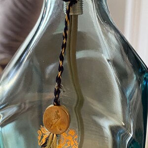 May include: A clear glass bottle with a gold seal and a black and gold cord. The seal has a gold embossed image of a man walking with a stick. The bottle has text that reads "By Appointment to Her Majesty The Queen Scotch Whisky Distillers John Walker & Sons Limited London".