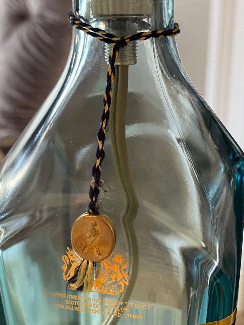 May include: A clear glass bottle with a gold seal and a black and gold cord. The seal has a gold embossed image of a man walking with a stick. The bottle has text that reads "By Appointment to Her Majesty The Queen Scotch Whisky Distillers John Walker & Sons Limited London".