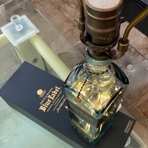 May include: A clear glass bottle lamp base with a gold-colored label that reads "Johnnie Walker Blue Label Blended Scotch Whisky" and a gold-colored tassel hanging from the top. The bottle is sitting on a blue box with the same label.