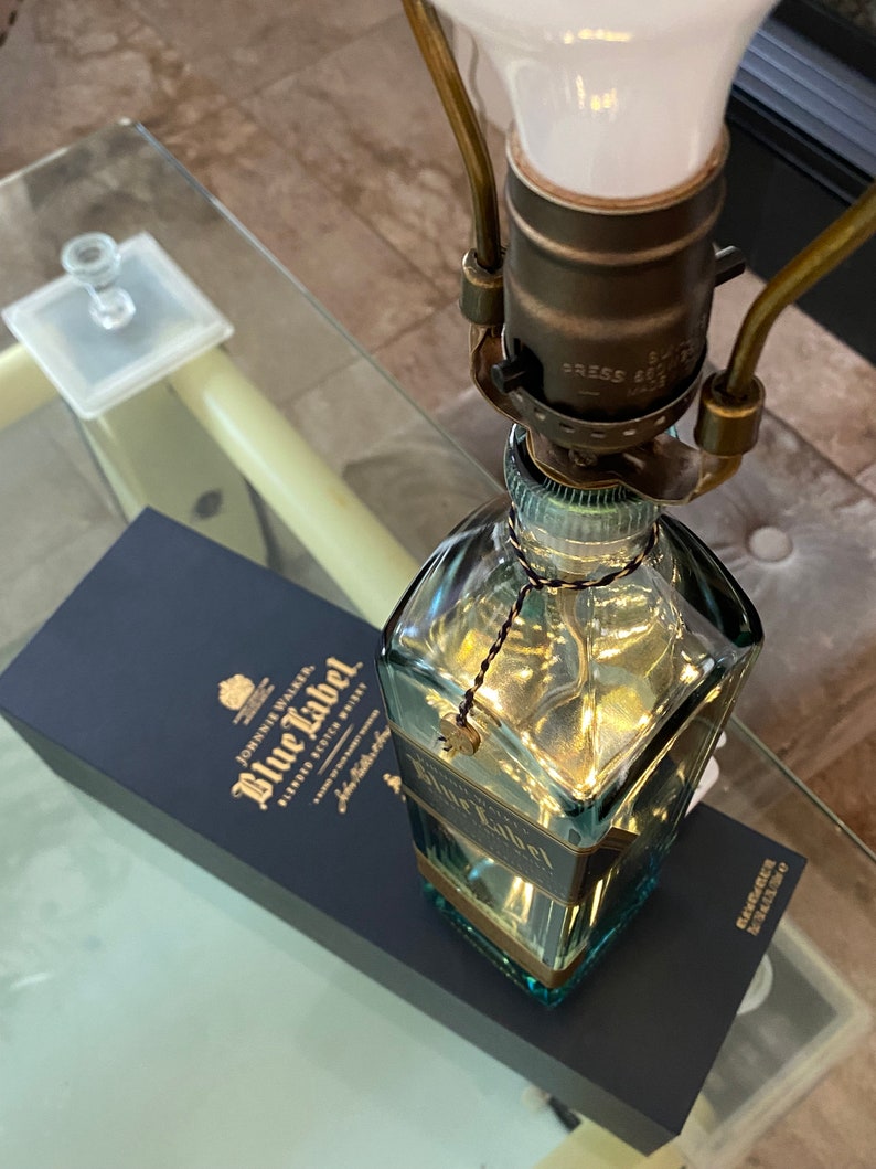 May include: A clear glass bottle lamp base with a gold-colored label that reads "Johnnie Walker Blue Label Blended Scotch Whisky" and a gold-colored tassel hanging from the top. The bottle is sitting on a blue box with the same label.