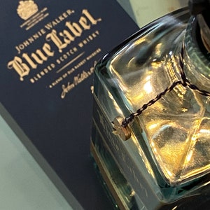 May include: A close-up of a clear glass bottle with a gold-colored cap and a black string tied around the neck. The bottle is sitting on top of a blue box with gold lettering that reads "Johnnie Walker Blue Label Blended Scotch Whisky".