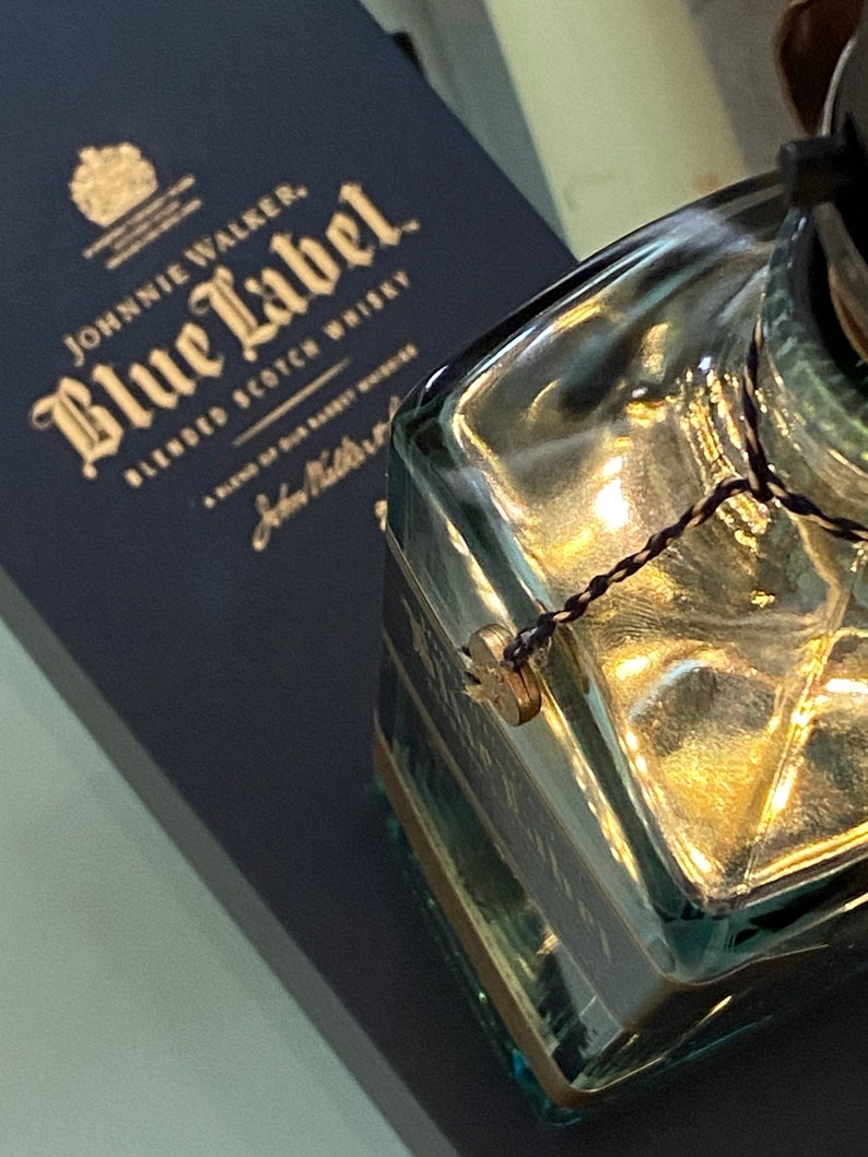 May include: A close-up of a clear glass bottle with a gold-colored cap and a black string tied around the neck. The bottle is sitting on top of a blue box with gold lettering that reads "Johnnie Walker Blue Label Blended Scotch Whisky".