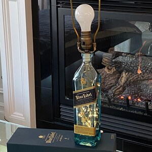 May include: A clear glass bottle with a gold label that reads "Johnnie Walker Blue Label Blended Scotch Whisky" is turned into a lamp with a gold metal lampshade and a white light bulb. The bottle is sitting on a black box with the same label.