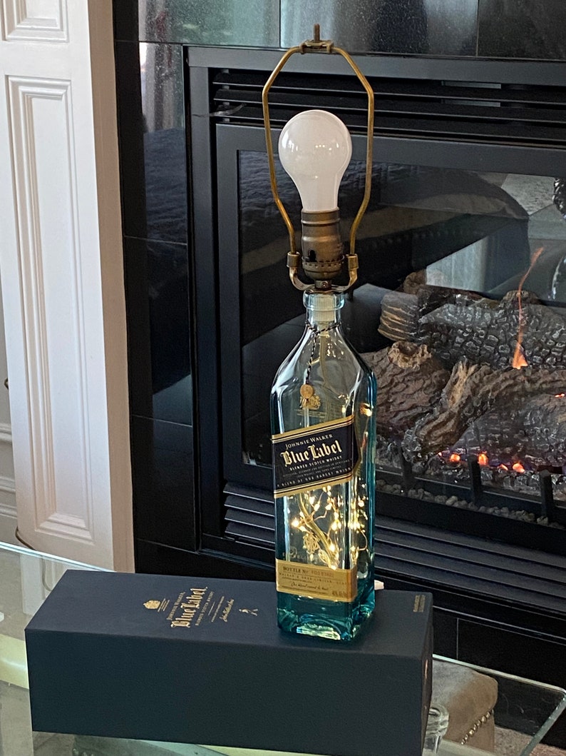 May include: A clear glass bottle with a gold label that reads "Johnnie Walker Blue Label Blended Scotch Whisky" is turned into a lamp with a gold metal lampshade and a white light bulb. The bottle is sitting on a black box with the same label.