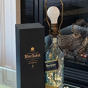 May include: A clear glass bottle with a gold label that reads "Johnnie Walker Blue Label Blended Scotch Whisky" is turned into a lamp with a white light bulb and a gold metal lampshade. The bottle is sitting on a glass table next to a black box with the same label.