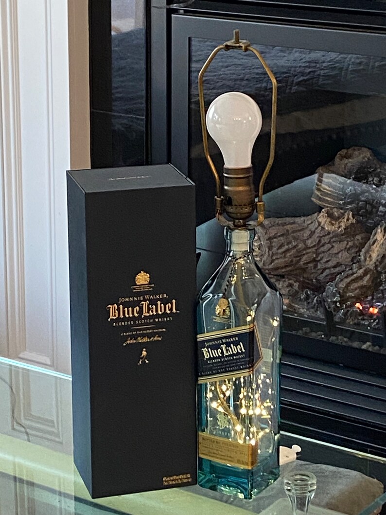 May include: A clear glass bottle with a gold label that reads "Johnnie Walker Blue Label Blended Scotch Whisky" is turned into a lamp with a white light bulb and a gold metal lampshade. The bottle is sitting on a glass table next to a black box with the same label.