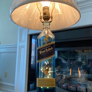May include: A repurposed Johnnie Walker Blue Label bottle turned into a table lamp with a white lampshade. The bottle is filled with white fairy lights and sits on a blue box with the Johnnie Walker Blue Label logo.