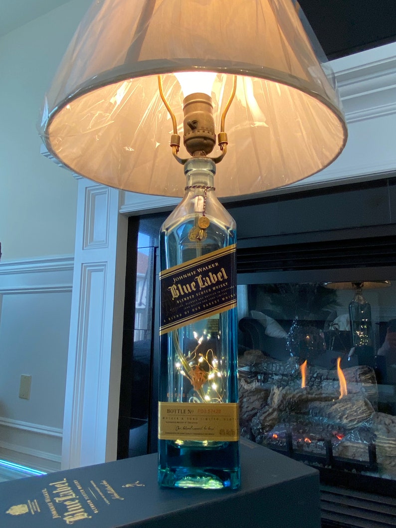 May include: A repurposed Johnnie Walker Blue Label bottle turned into a table lamp with a white lampshade. The bottle is filled with white fairy lights and sits on a blue box with the Johnnie Walker Blue Label logo.
