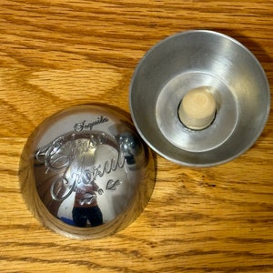 May include: Silver bottle stopper with a round top and a small, round, wooden stopper inside. The top of the bottle stopper has the words "Tequila Azul" engraved on it.