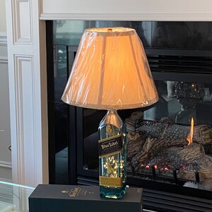 May include: A repurposed bottle of Johnnie Walker Blue Label Scotch Whisky turned into a table lamp with a white lampshade. The bottle is filled with white fairy lights and has a blue liquid inside. The lamp is sitting on a black box with the Johnnie Walker Blue Label logo.