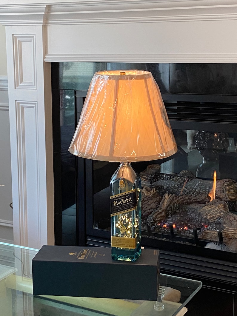 May include: A repurposed bottle of Johnnie Walker Blue Label Scotch Whisky turned into a table lamp with a white lampshade. The bottle is filled with white fairy lights and has a blue liquid inside. The lamp is sitting on a black box with the Johnnie Walker Blue Label logo.