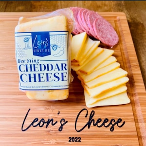 May include: A block of Bee Sting Cheddar Cheese wrapped in plastic on a wooden cutting board with slices of salami and cheddar cheese. The cheese is labeled "Lein's Cheese Est. 2022 Bee Sting Cheddar Cheese". The cutting board is labeled "Leon's Cheese 2022".