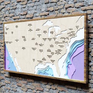May include: A framed wooden map of the USA, displayed on a brick wall. The map, with a gold frame, shows detailed laser-cut coastlines and state outlines, with city names. The ocean depths are represented with layers of blue and purple.