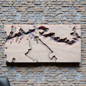 May include: A wooden relief map of a geographical region, likely a coastline, with layered contours. The map is light-colored wood with blue and red accents, mounted on a brick wall. The design highlights the topography.