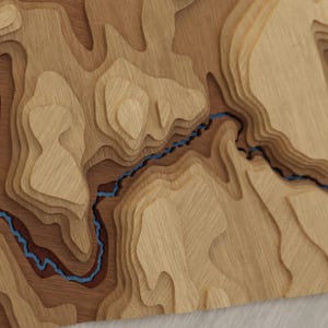 May include: A multi-layered wooden topographic map featuring varying shades of natural wood grain. The design includes raised contour levels that create a three-dimensional effect, with a thin, winding blue river carved through the center.