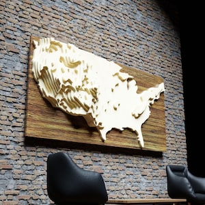 May include: A wooden wall art piece depicting a 3D relief map of the United States. The map is light-colored and mounted on a rectangular, dark-stained wooden board. The artwork is displayed against a brick wall.