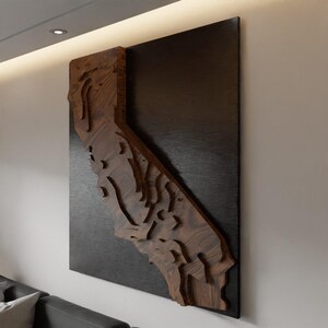 May include: A wooden wall art piece depicting the state of California. The artwork features a detailed, layered design in a dark wood tone, mounted on a dark, rectangular background. The piece is displayed on a white wall, showcasing the state's topography.