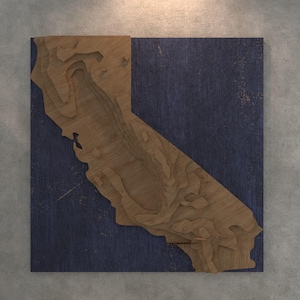 May include: A wooden wall art piece featuring a detailed, layered map of California in natural wood tones. The map is set against a dark blue background, creating a striking contrast. The artwork is square and mounted on a gray wall.