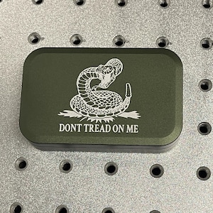 May include: An olive green rectangular box with rounded corners. The box features a white graphic of a coiled snake above the words "DONT TREAD ON ME". The box is on a silver surface with evenly spaced holes.