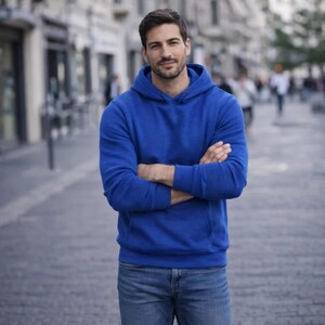 May include: A person wearing a royal blue hoodie with a front pocket and blue jeans. The person is standing outside with arms crossed. The background is a blurred city street.