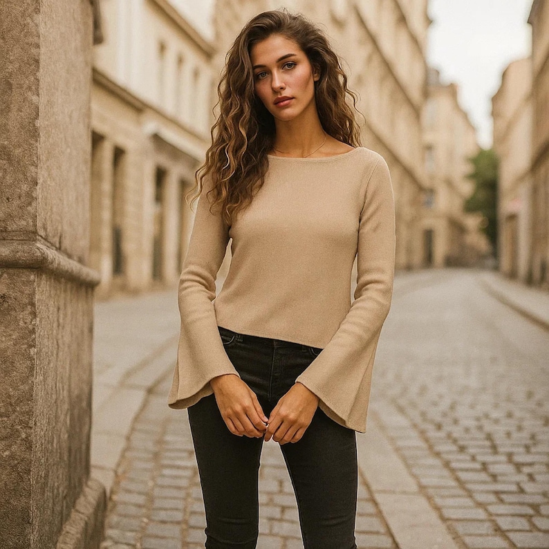 May include: A beige long-sleeve sweater with flared sleeves and a boat neck. The sweater is paired with black jeans. The background shows a cobblestone street and buildings.