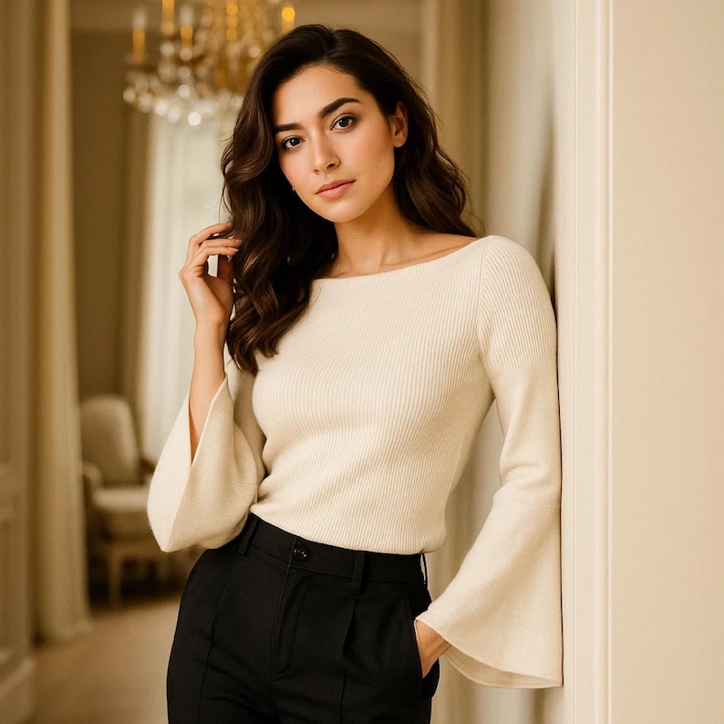 May include: Cream-colored ribbed knit sweater with a boat neck and flared sleeves, paired with black trousers. The sweater's design features a fitted bodice and long sleeves that widen towards the wrist. The outfit is simple and elegant.