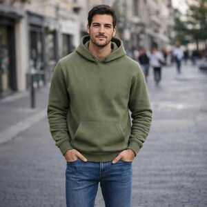 May include: An olive green hooded sweatshirt with a kangaroo pocket. The sweatshirt is paired with blue jeans. The person is standing on a paved street with buildings in the background.