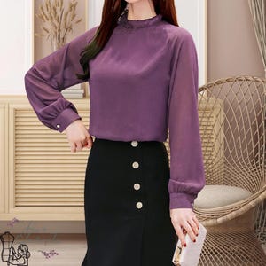 May include: A purple long-sleeve blouse with a ruffled neckline and buttoned cuffs. The blouse is paired with a black skirt featuring decorative buttons. The outfit is suitable for a professional or semi-formal setting.