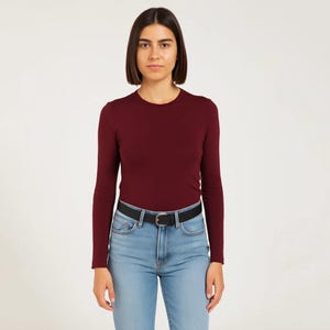 May include: A long-sleeved burgundy shirt with a crew neck, paired with light blue jeans and a black belt. The shirt is a solid color, and the jeans have a classic five-pocket design. The belt has a silver buckle.