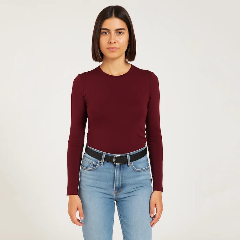 May include: A long-sleeved burgundy shirt with a crew neck, paired with light blue jeans and a black belt. The shirt is a solid color, and the jeans have a classic five-pocket design. The belt has a silver buckle.