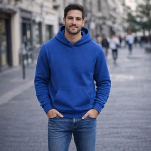 May include: A royal blue hoodie with a kangaroo pocket and a drawstring hood. The hoodie is paired with blue jeans. The person is standing on a paved street with blurred background elements.