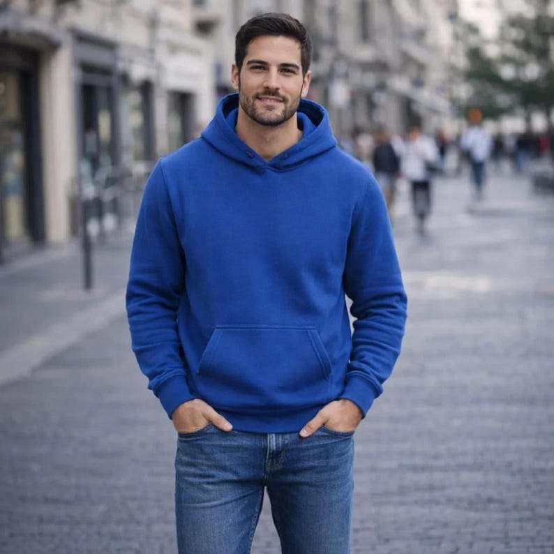 May include: A royal blue hoodie with a kangaroo pocket and a drawstring hood. The hoodie is paired with blue jeans. The person is standing on a paved street with blurred background elements.