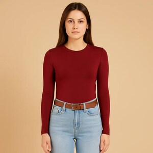 May include: A long-sleeved, burgundy crew neck shirt paired with light blue jeans and a brown leather belt. The belt has a silver buckle. The outfit is simple and casual.