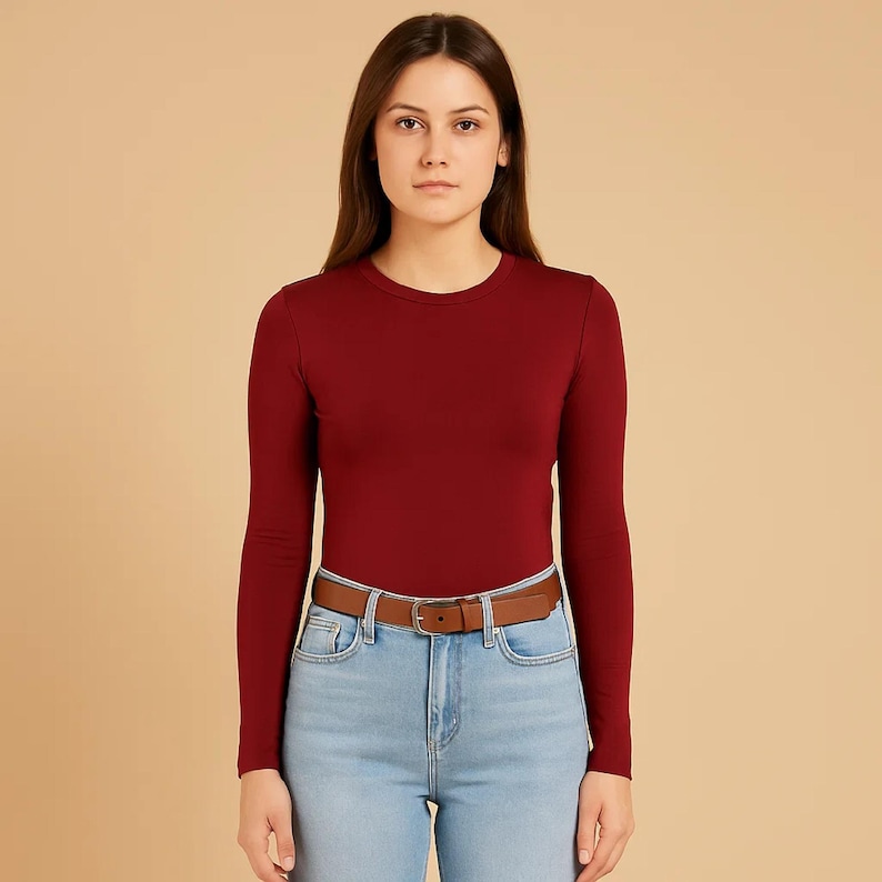 May include: A long-sleeved, burgundy crew neck shirt paired with light blue jeans and a brown leather belt. The belt has a silver buckle. The outfit is simple and casual.