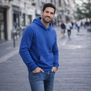 May include: A person wearing a royal blue hoodie with a front pocket and blue jeans. The hoodie has a drawstring hood and long sleeves. The person is smiling and standing on a city street.