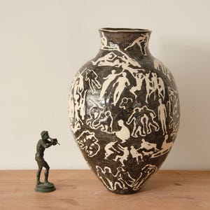 Może przedstawiać: Duży, ciemnobrązowy wazon ceramiczny z białą glazurą przedstawiającą abstrakcyjne figury. Wazon ma zaokrąglone ciało i wąską szyjkę.
