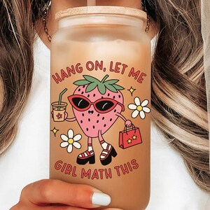 May include: A clear glass reusable water bottle with a bamboo lid and a strawberry character graphic. The graphic says "Hang on, let me girl math this".