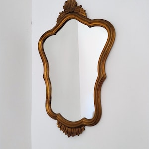 Könnte beinhalten: Ein goldfarbener Spiegel mit geschwungenem Rahmen und dekorativen Elementen. Das Design ist antik und elegant. Der Spiegel hängt an einer weißen Wand und ist ein Blickfang.